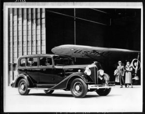 Packard Co. file photograph of a 1934 Packard seven-eights right side view, couple and airplane hangar in background, airplane wing reads "U.S. Mail." Inscribed on photo back: "Packard 1100 eight, eleventh series, 8-cylinder, 120-horsepower, 129-inch wheelbase, 5-person sedan (body type #703), fitted with Packard deluxe emblem radiator cap."