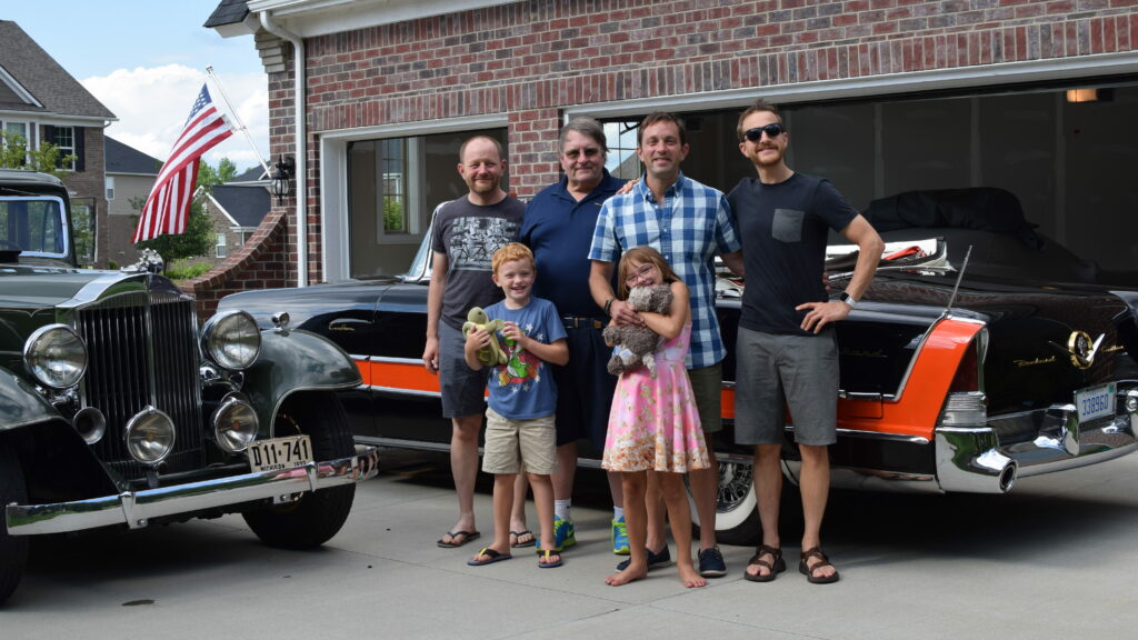 Dave, family, and two Packards on a driveway next to a garage, with an American flag flying in the background