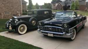 Two black Packards, each from a different decade, parked on a driveway in a neighborhood