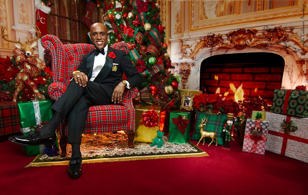 Carlossee’ sitting in a chair in front of a decorated Christmas tree with presents underneath, next to a fireplace